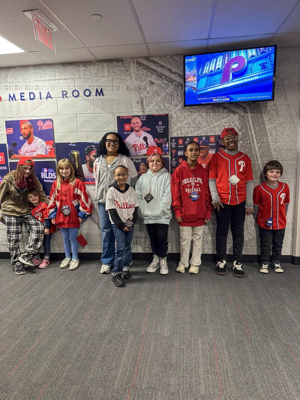 A group of young PALCS students wearing Phillies apparel posing together outside the Citizens Bank Park Media Room.