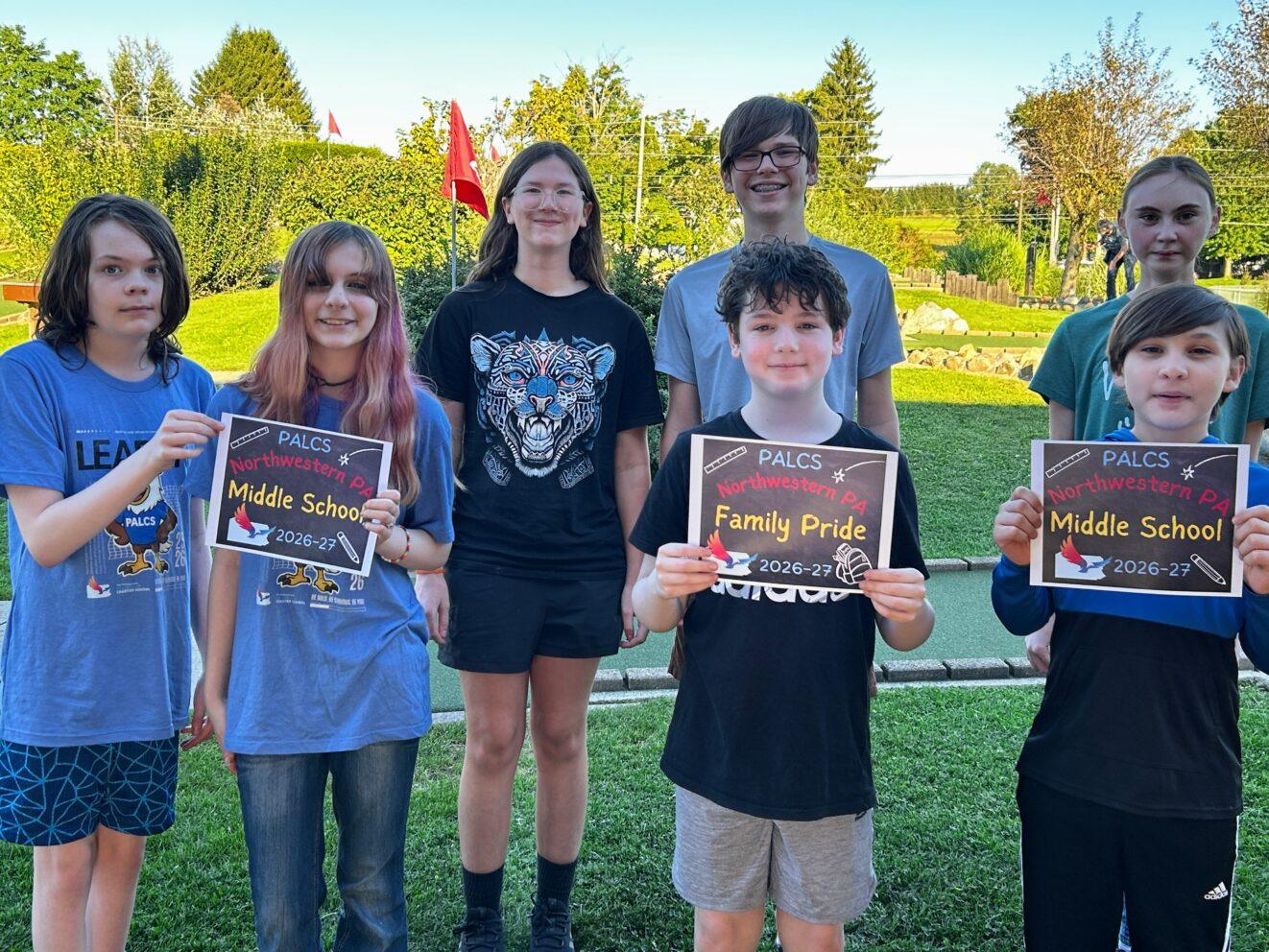 A group of PALCS students and parents posing outdoors together while holding "PALCS Middle School 2026-27" and "PALCS Family Pride" signs.