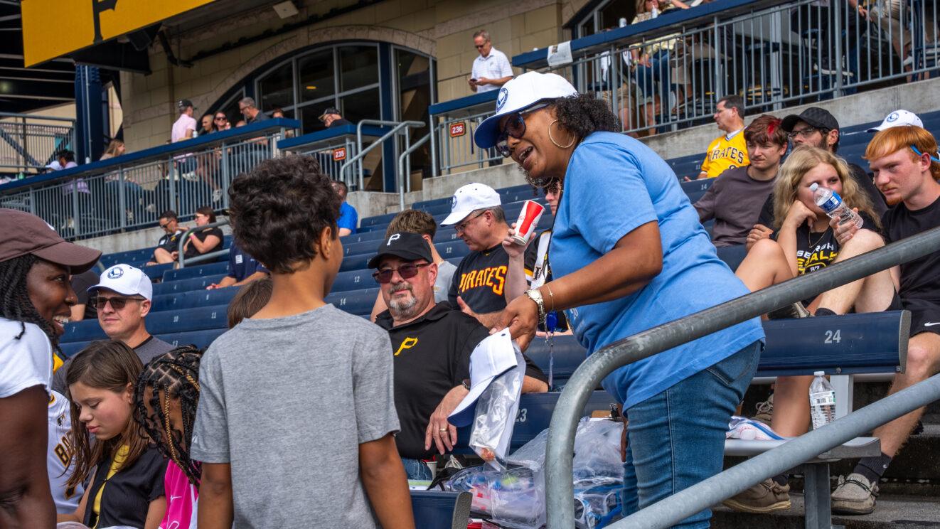 A PALCS employee wearing a blue shirt speaks to a PALCS students who is wearing a gray shirt. This interaction in taking place in the stands at a Pittsburgh Pirates baseball game, and there are other people in the stands.