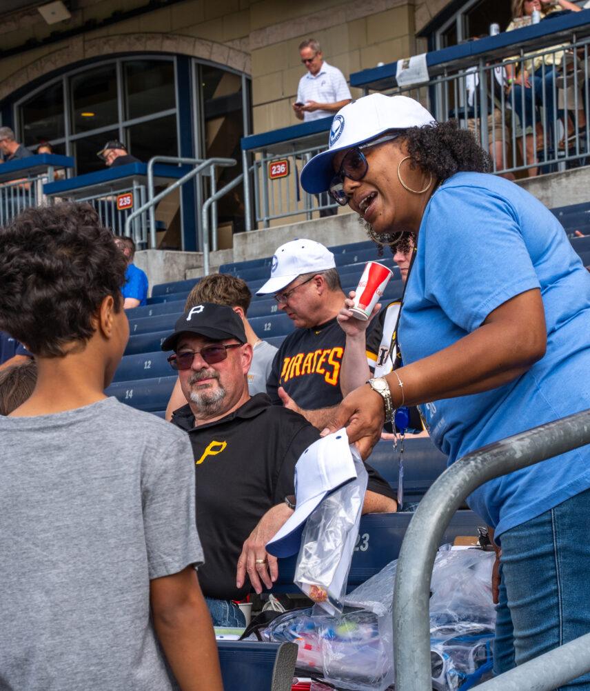 A PALCS employee wearing a blue shirt speaks to a PALCS students who is wearing a gray shirt. This interaction in taking place in the stands at a Pittsburgh Pirates baseball game, and there are other people in the stands.
