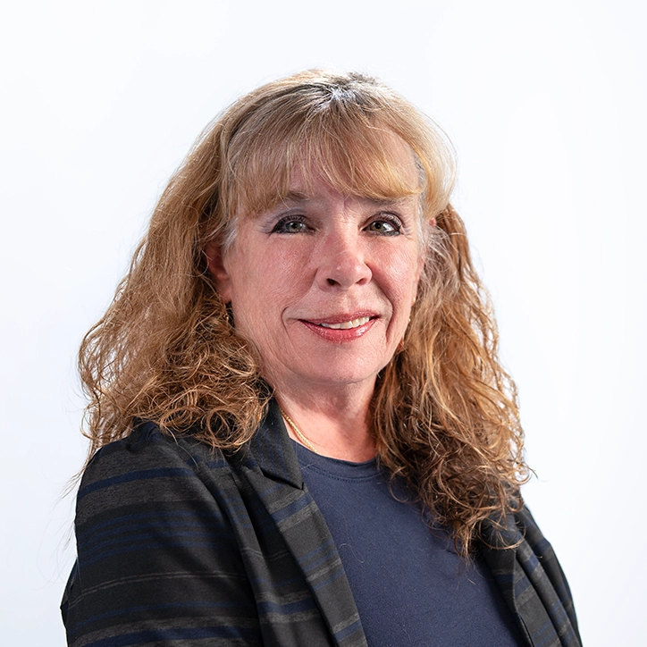 A PALCS staff member with curly auburn hair and a dark striped jacket smiles for a portrait against a light background.